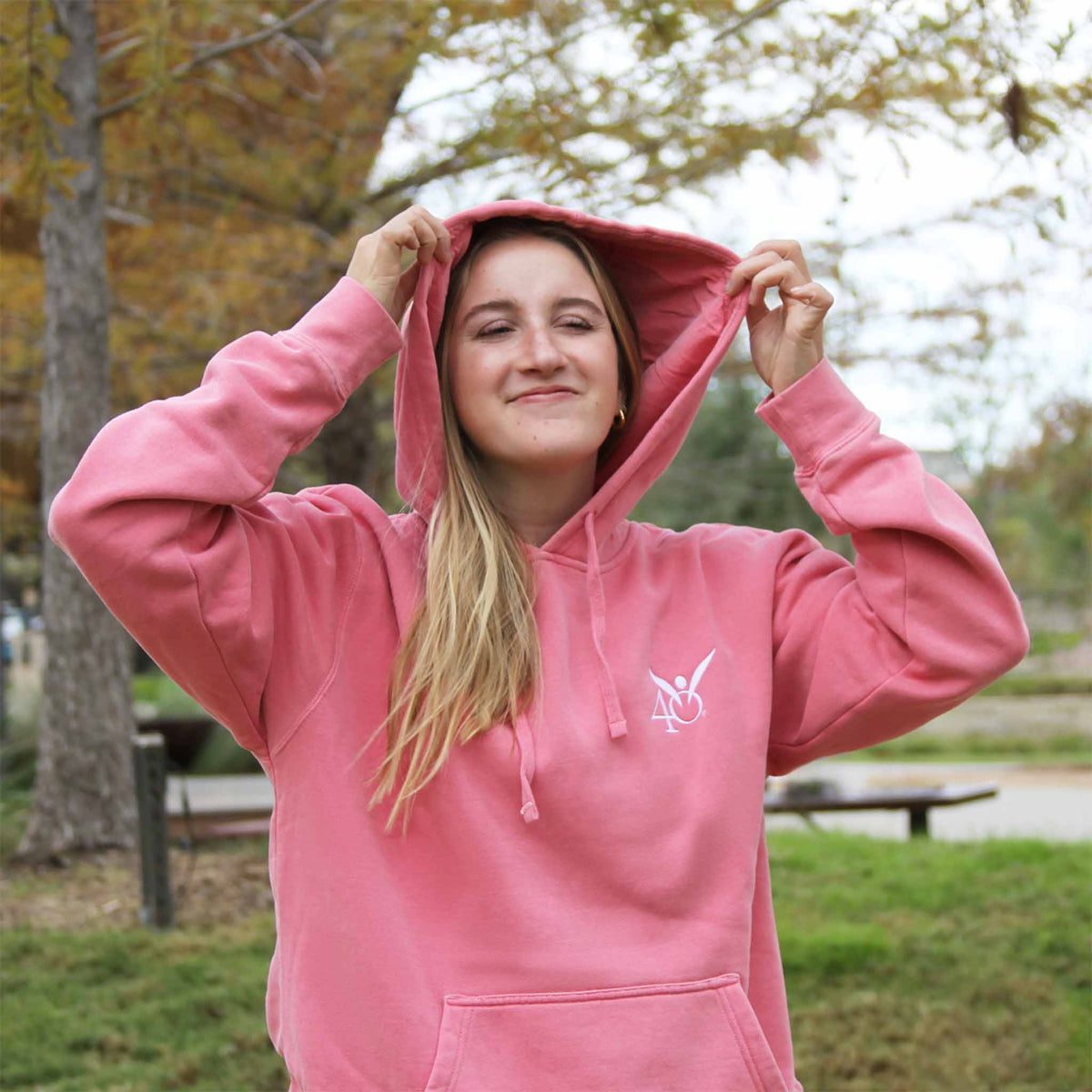 A woman wearing a pink hoodie with an embroidered angel symbol on the front and the hood is up, showing the logo on the front of the hoodie.