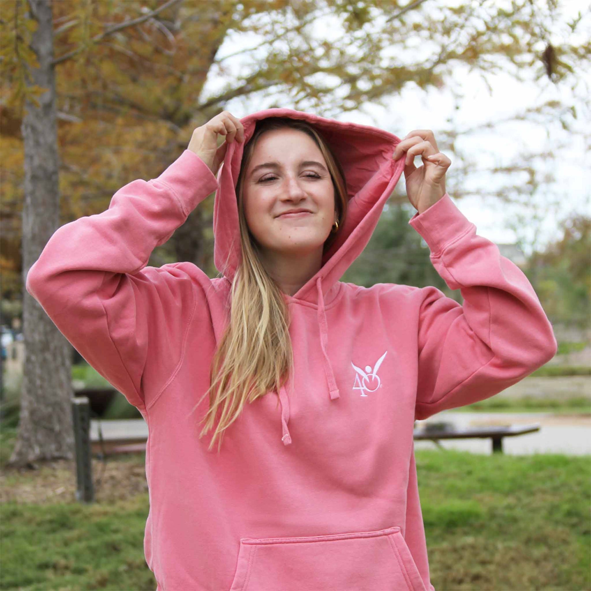 A woman wearing a pink hoodie with an embroidered angel symbol on the front and the hood is up, showing the logo on the front of the hoodie.
