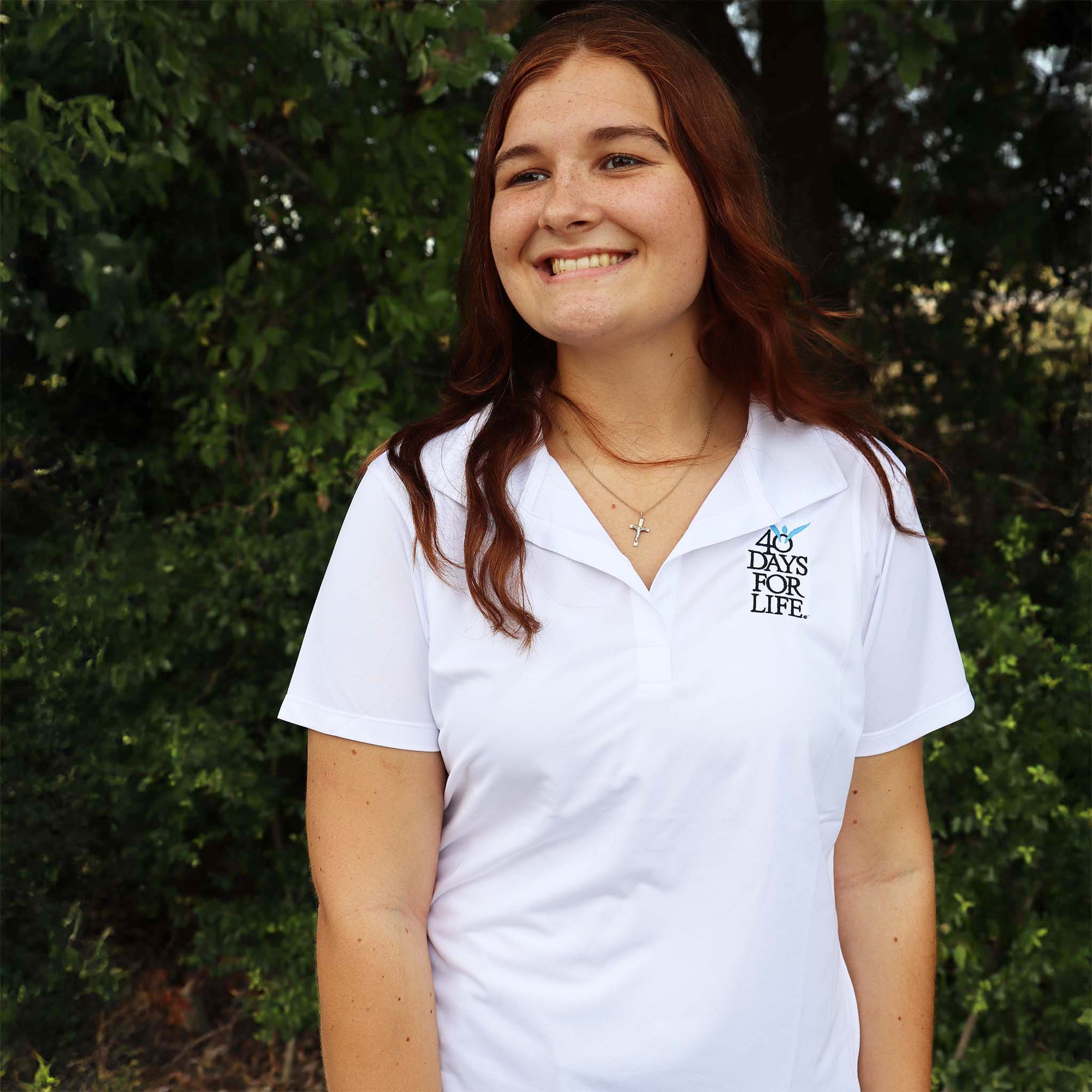 Female wearing a white polo shirt with a logo in front of green foliage
