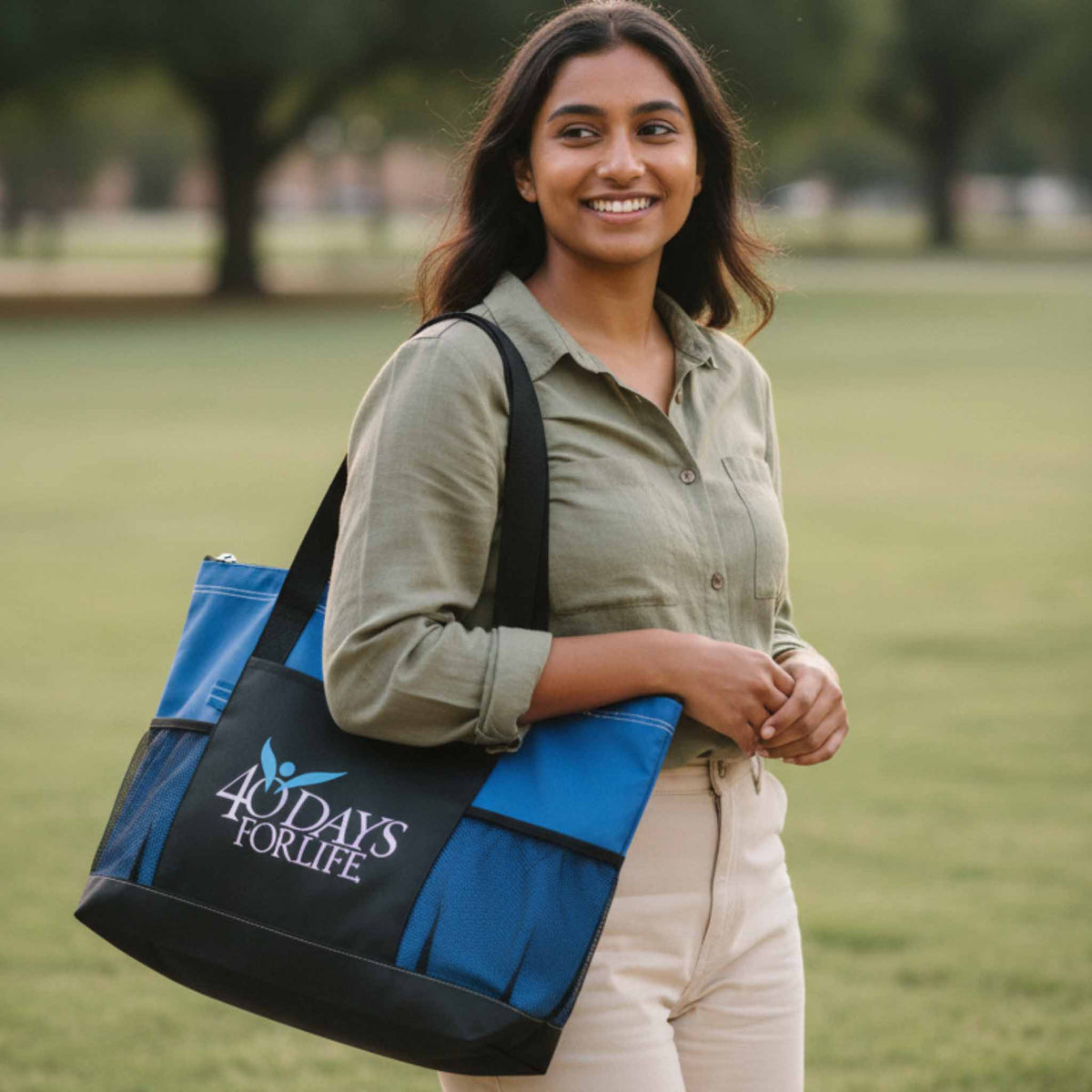 A woman in a green shirt and beige pants stands in a grassy park holding a blue and black tote bag with the logo '40 DAYS FOR LIFE'.
