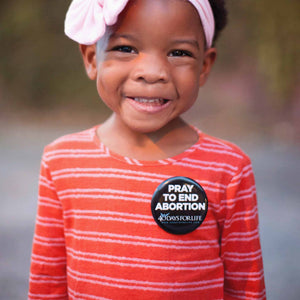 A young child wearing a pink bow headband and a red and white striped shirt, with a black button on the chest that reads 'PRAY TO END ABORTION 40 Days For Life'.